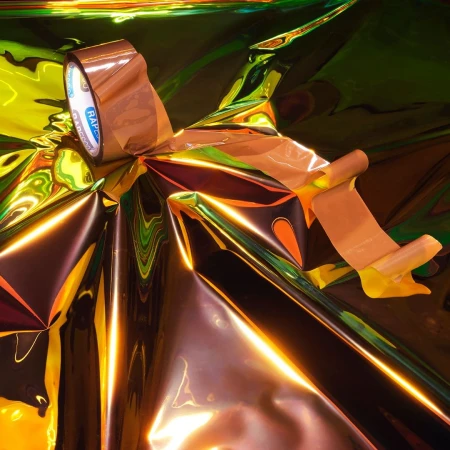 Shiny iridescent material is partially covered with copper-toned tape, partially unrolled to reveal the shiny yellow material underneath.