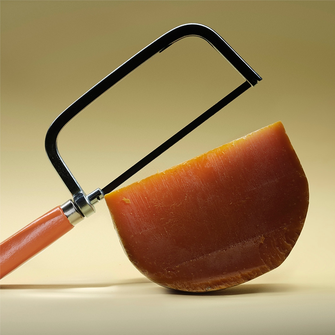 A semi-circular wedge of orange cheese is being “cut” by a small, handheld, chrome, u-shaped saw on a yellow surface and background.