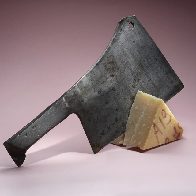 Metal cleaver and a wedge of cheese on a pink surface, studio lit. Close-up shot featuring textures and contrasting tones.
