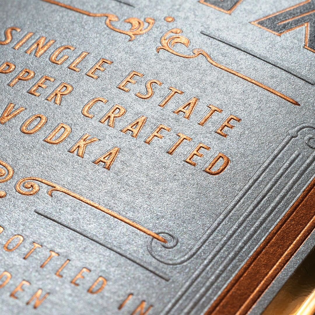 Close-up product shot of premium vodka label with copper-colored embossed text and decorative borders on a textured gray surface.
