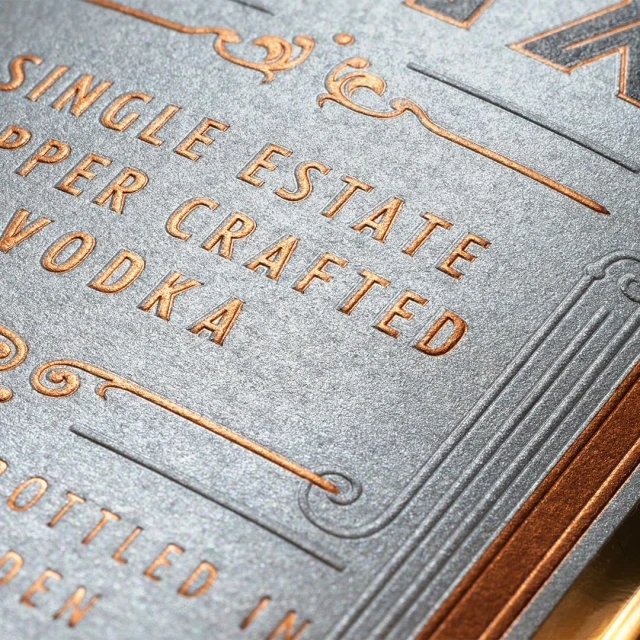 Close-up product shot of premium vodka label with copper-colored embossed text and decorative borders on a textured gray surface.