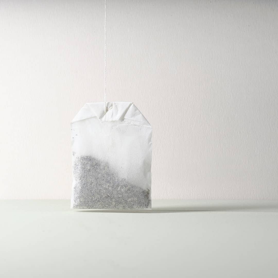Single tea bag hanging from a string against a plain white background, casting a soft shadow on the surface below in bright studio lighting.