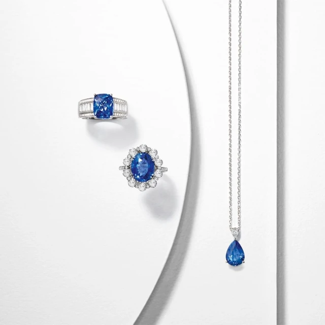 Close-up shot of two rings and a necklace with blue gems and diamond accents displayed on a white surface with soft, diffused lighting.