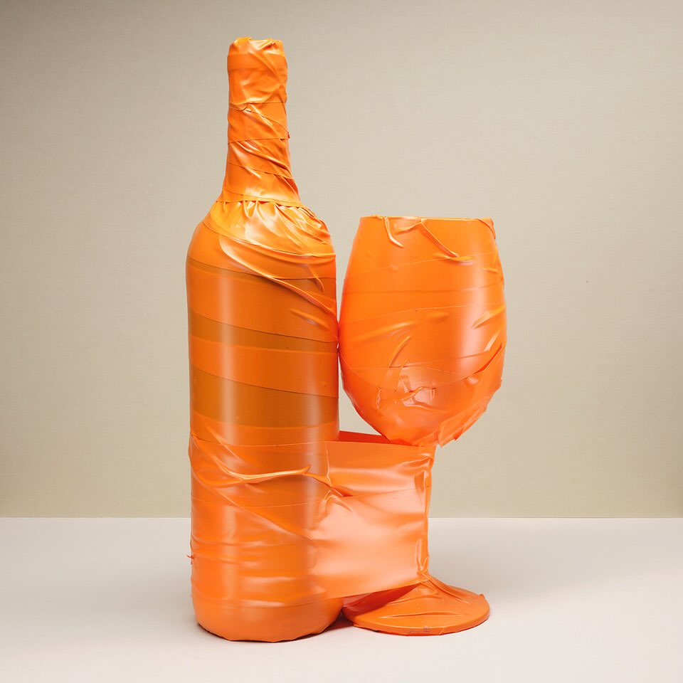 Abstract still life featuring an orange wine bottle and glass wrapped in tape on a white surface against a light beige background.