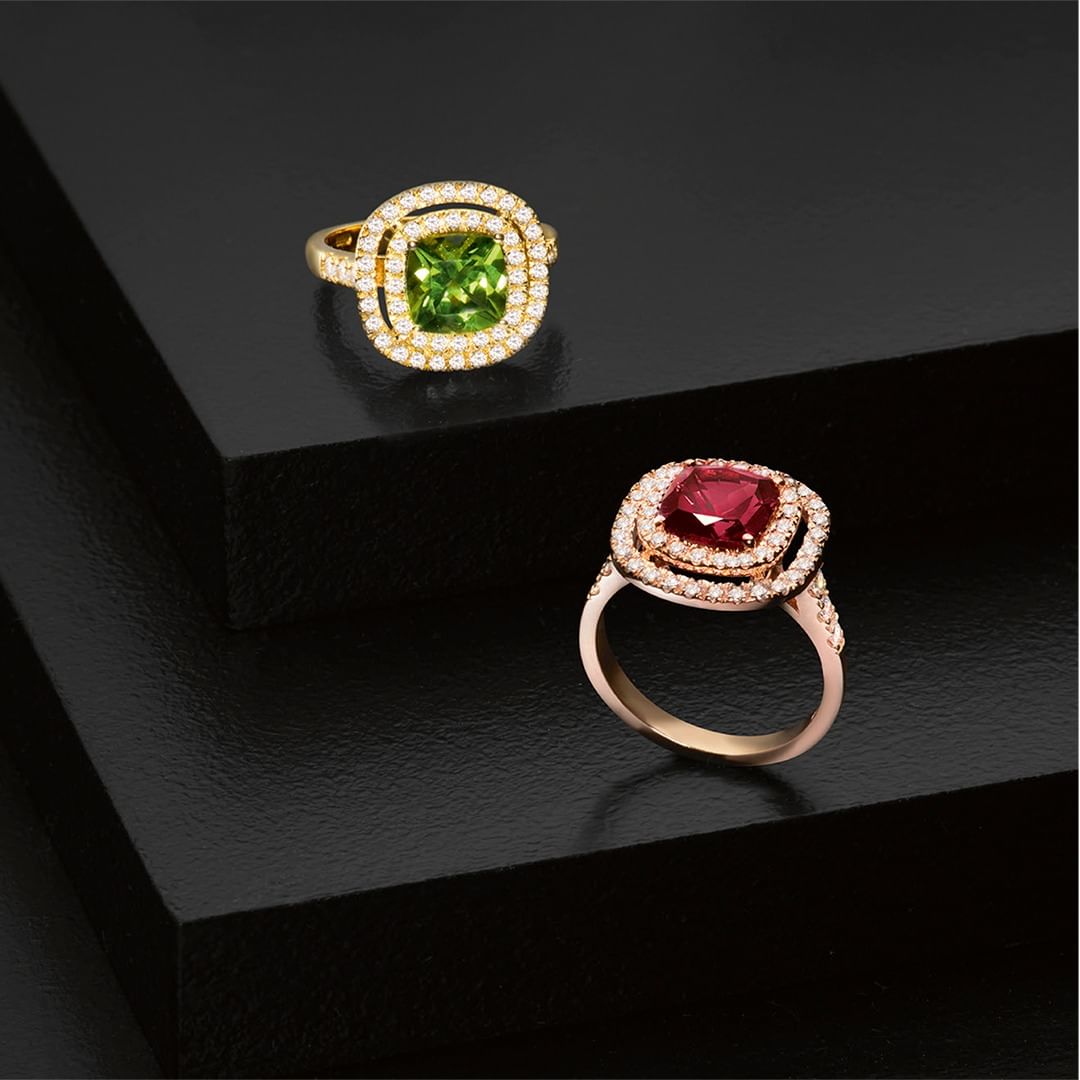 Two gold rings with square gemstones and diamond halos are displayed on a sleek black surface in a studio jewelry shot.