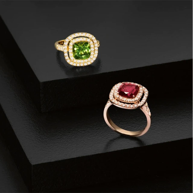 Two gold rings with square gemstones and diamond halos are displayed on a sleek black surface in a studio jewelry shot.