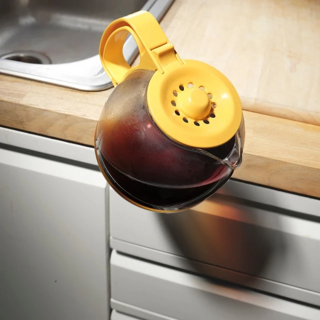Clear glass pitcher filled with dark liquid on light wood counter next to a stainless steel sink and white cabinets.