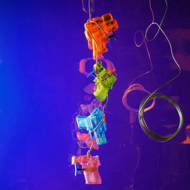 Four translucent toy water guns are suspended on wire against a mottled blue and pink background, creating a playful and colorful scene.
