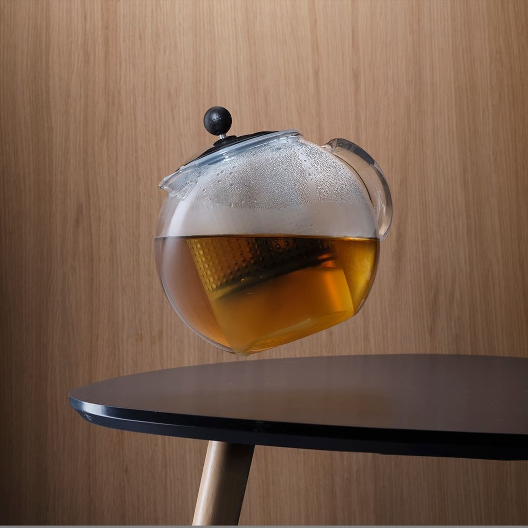 Glass teapot with amber liquid sits on a black table against a wood panel backdrop in a balanced composition.