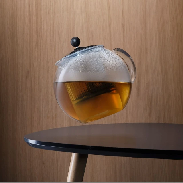 Glass teapot with amber liquid sits on a black table against a wood panel backdrop in a balanced composition.
