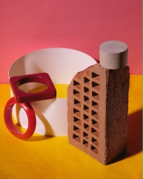 Still life photography featuring a perforated red brick, a grey concrete cylinder, and red and burgundy geometric bangles on a yellow and pink surface.