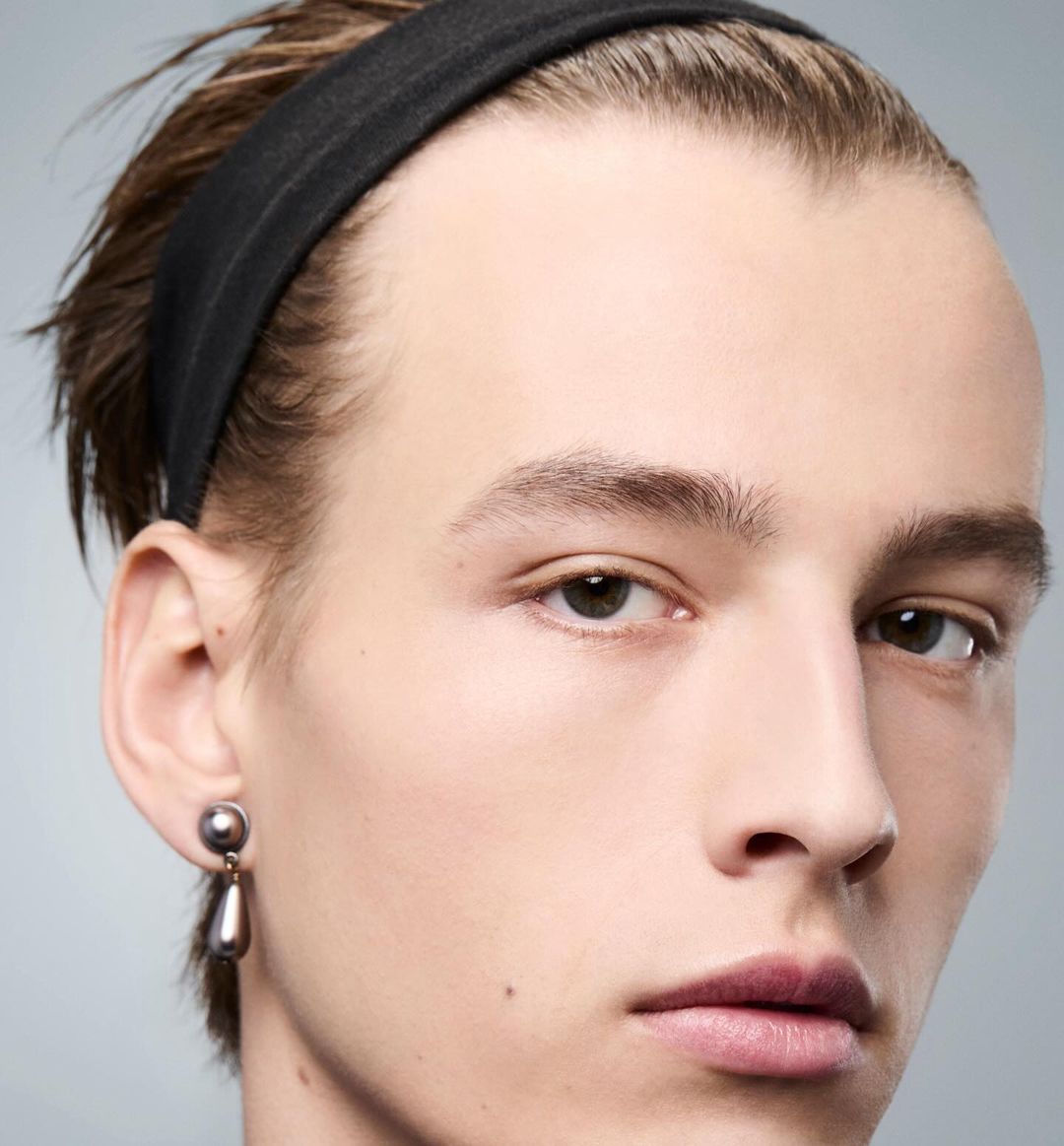 Close-up studio portrait of a young adult male with neutral expression wearing a black headband and a tear-drop earring.