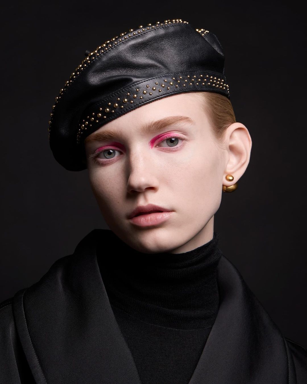 Close-up portrait of a woman wearing a black leather beret with gold studs and a black turtleneck, set against a dark background.