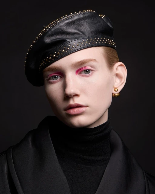 Close-up portrait of a woman wearing a black leather beret with gold studs and a black turtleneck, set against a dark background.