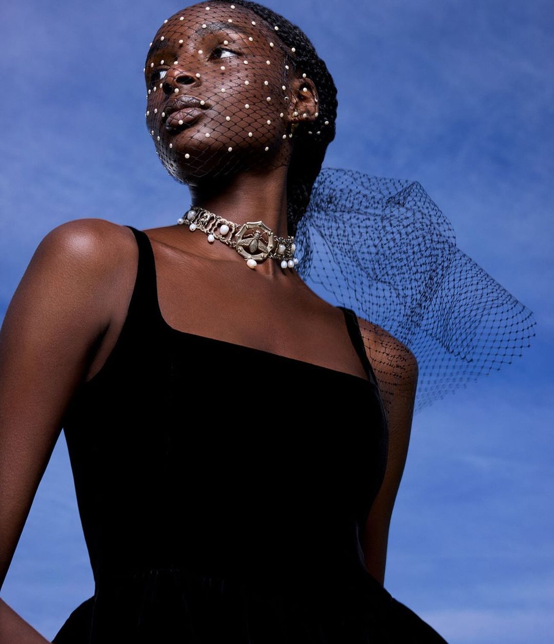 Close-up fashion portrait of a Black female model wearing a black dress, pearl choker, and a beaded black veil against a blue sky background.