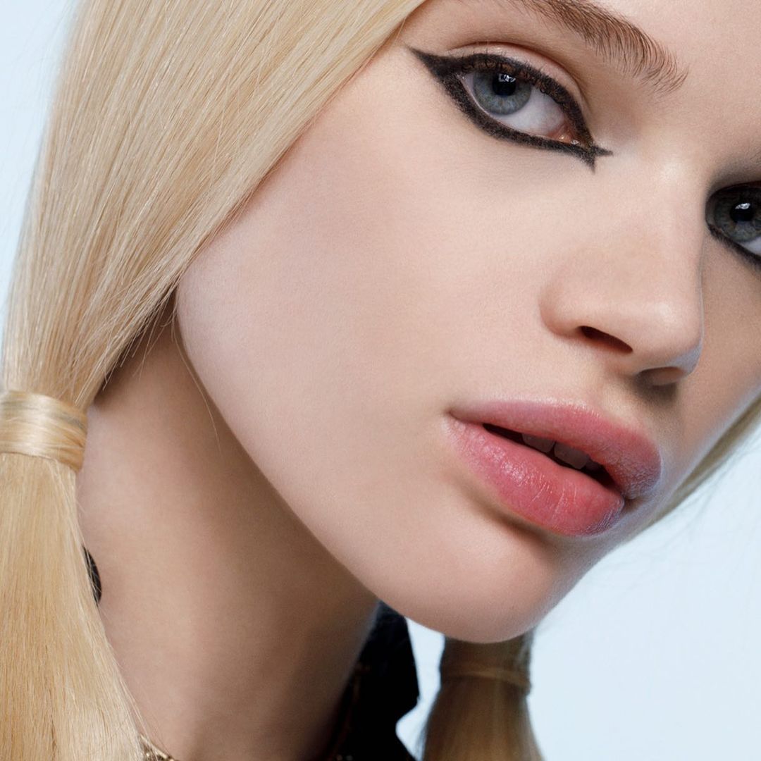 Close-up studio shot of a young adult female with blonde hair in pigtails, wearing eyeliner and pink lipstick, against a blue background.