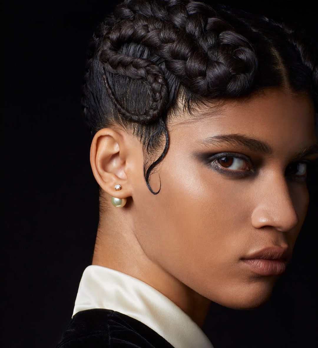 Close-up portrait of a young adult woman with braided hair, smokey eyeshadow, a white collared shirt, and pearl earrings against a black background.