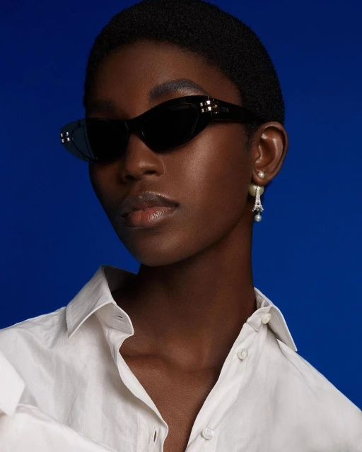 Close-up of a Black woman wearing sunglasses, a white shirt and Eiffel Tower earrings, against a blue background.