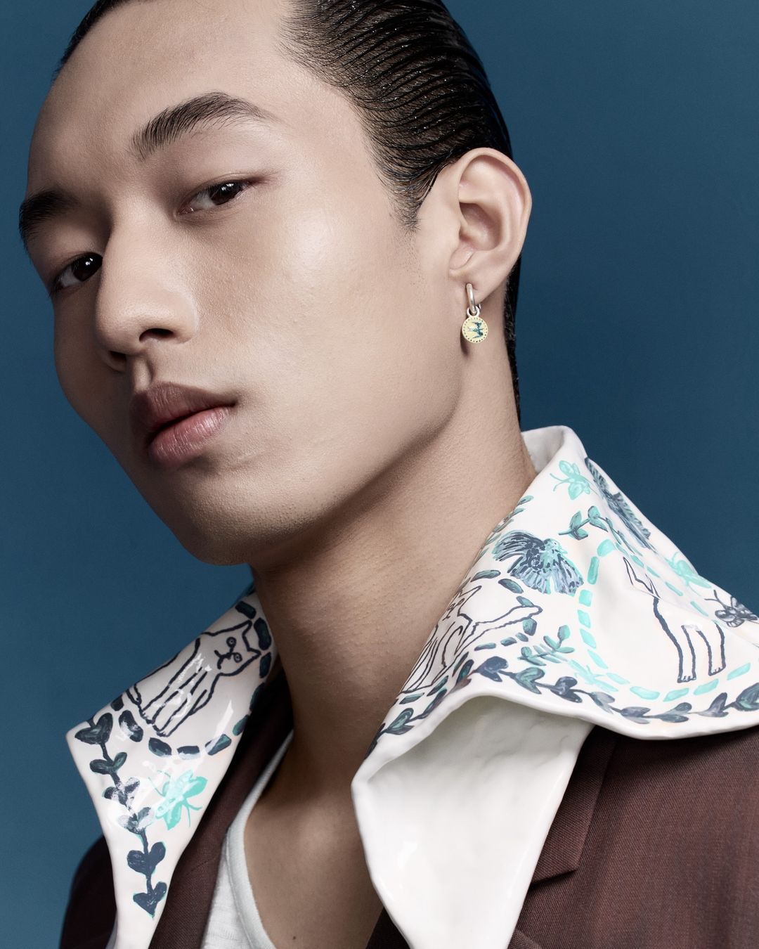Close-up portrait of an East Asian man wearing a patterned white collar and brown jacket against a solid blue background.