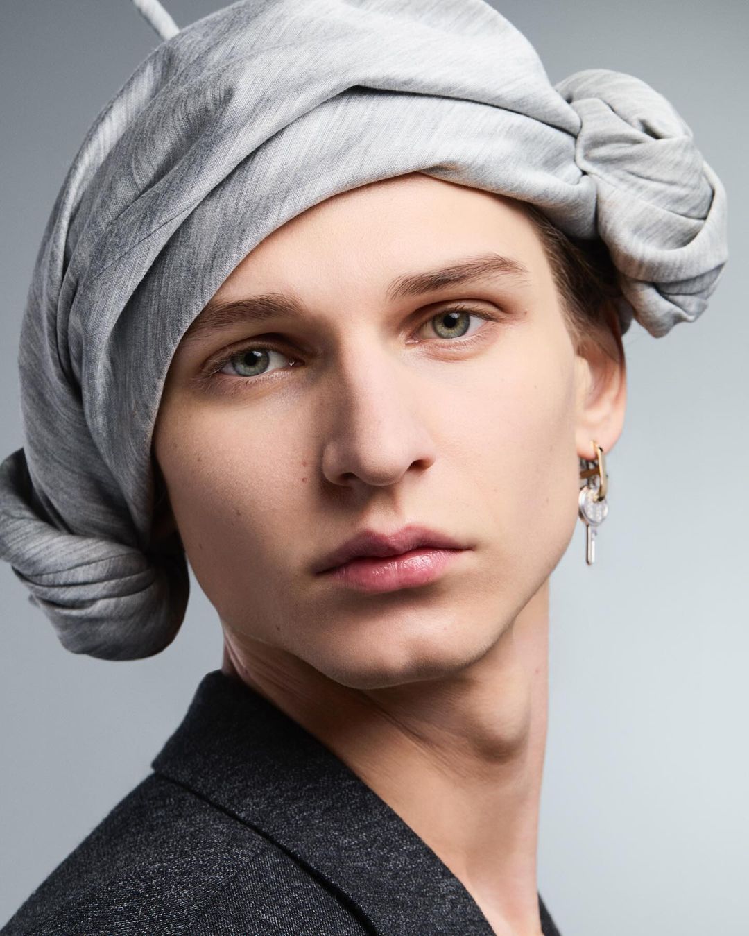 Close-up of a young adult male wearing a gray head wrap and dark blazer, set against a neutral gray background. Fashion editorial style.