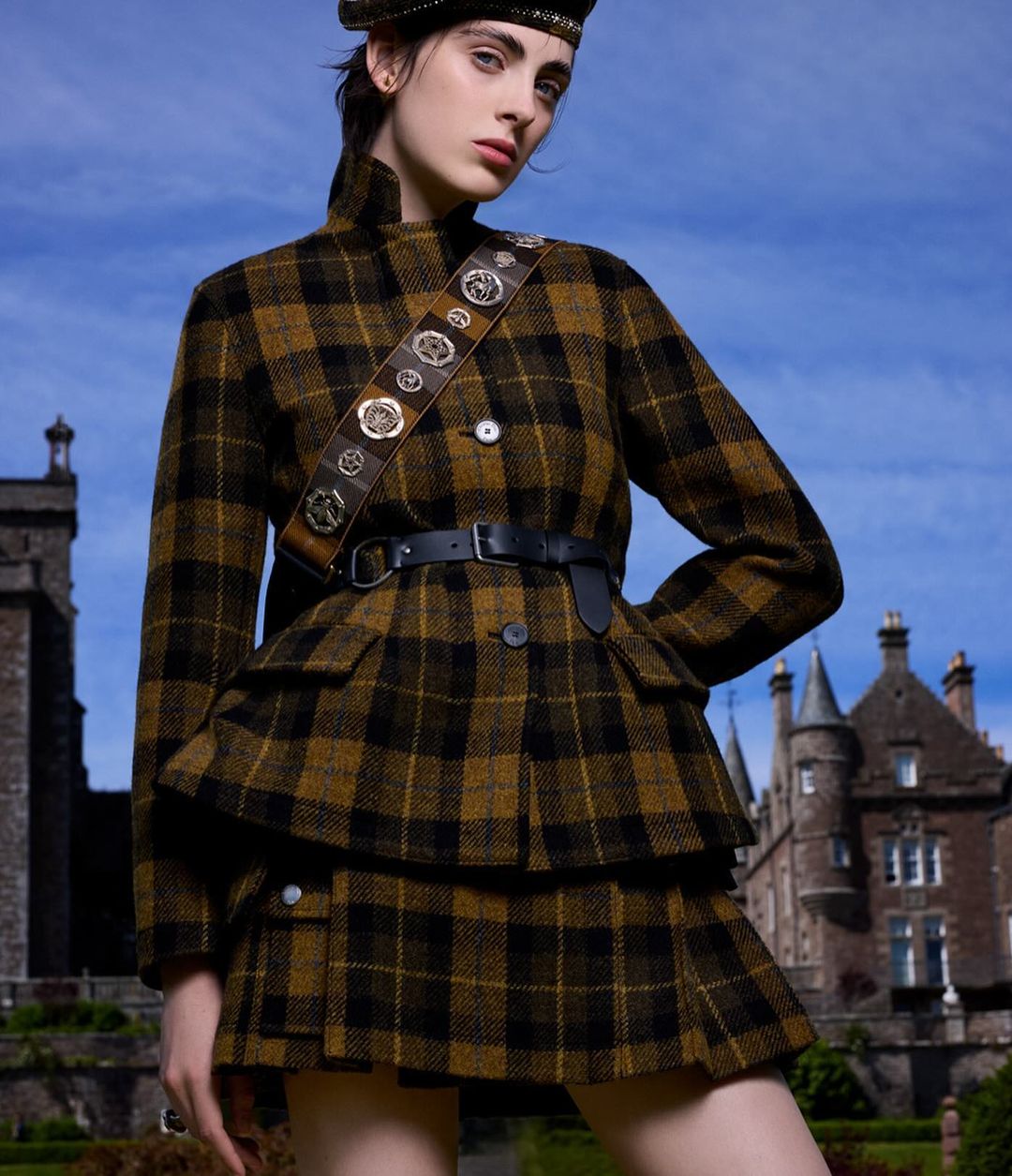 A woman wearing a brown plaid skirt and jacket with a matching beret, against a backdrop of a blue sky and a historic castle.