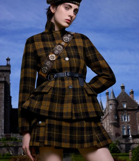 A woman wearing a brown plaid skirt and jacket with a matching beret, against a backdrop of a blue sky and a historic castle.