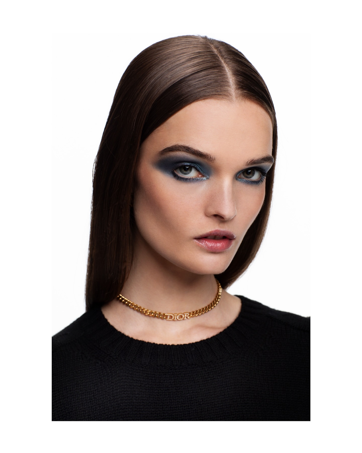 Close-up shot of a woman with dark hair and smoky blue eyeshadow wearing a gold Dior choker against a white backdrop.