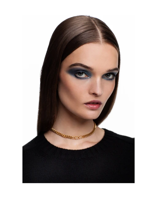 Close-up shot of a woman with dark hair and smoky blue eyeshadow wearing a gold Dior choker against a white backdrop.