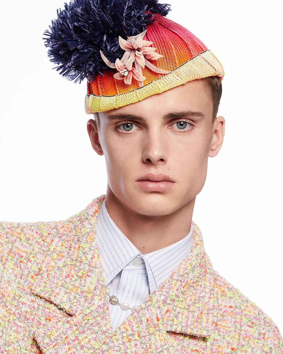 A fashionable young adult poses in a colorful tweed jacket and a knitted hat with a large pompom and floral embellishment against a white backdrop.