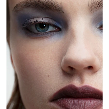 Close-up portrait of a woman's face, featuring blue eyeshadow, a mole near her nose, and dark burgundy lipstick.