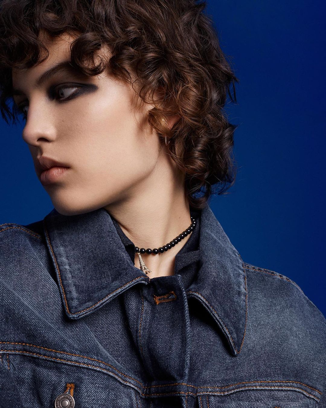 A young woman with dark eye makeup wearing a denim jacket and a black beaded necklace poses against a royal blue backdrop.