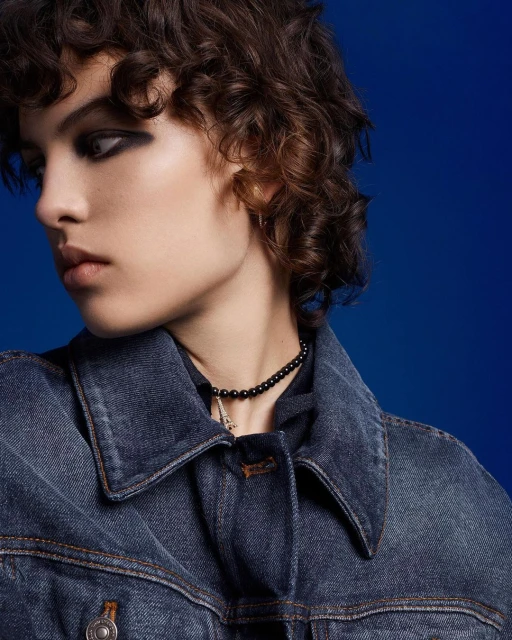 A young woman with dark eye makeup wearing a denim jacket and a black beaded necklace poses against a royal blue backdrop.