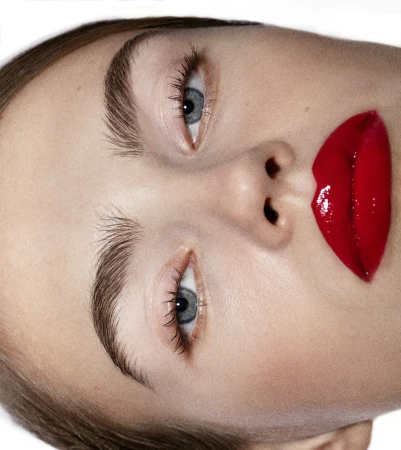 Extreme close-up portrait of a woman with blue eyes, groomed eyebrows, and glossy red lipstick against a plain white background.