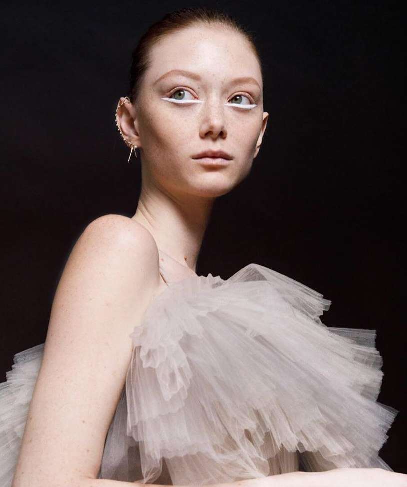 Close-up portrait of a fair-skinned adult woman with red hair, white eyeliner, statement ear jewelry, and a layered white tulle dress against black.