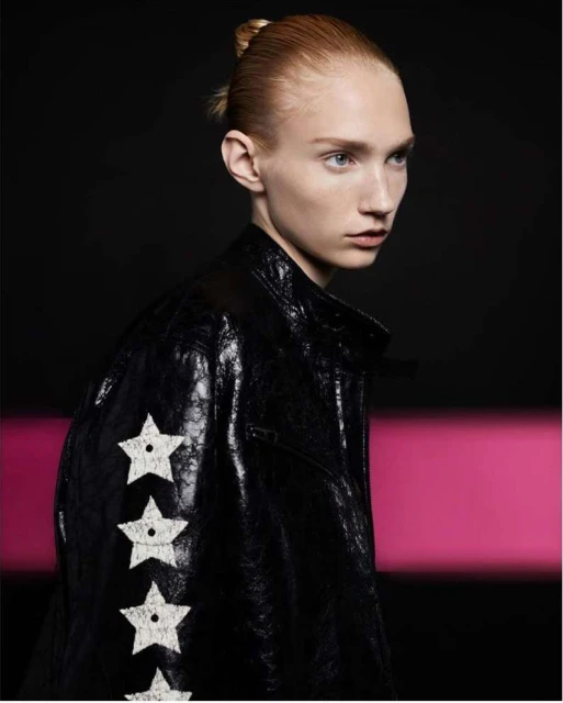 Adult fashion model poses indoors wearing a black leather jacket with white star pattern and wearing a top knot hairstyle.