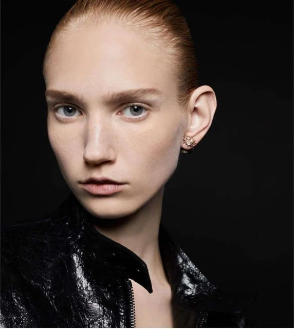Close-up studio shot of a fair-skinned young adult female with light hair, wearing a black leather jacket and earrings against a dark backdrop.