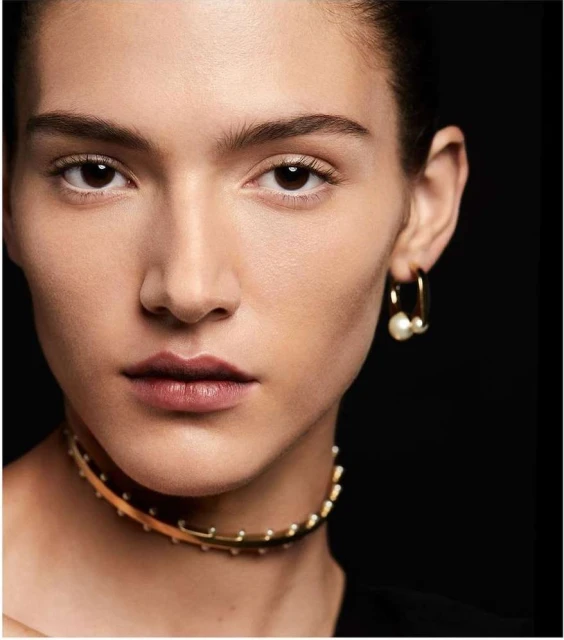 Close-up studio shot of a young adult female with dark hair wearing a gold choker necklace and pearl earrings against a black background.