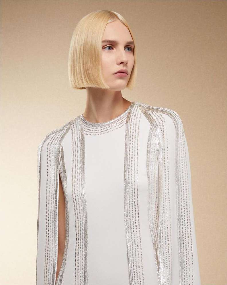 Blonde woman with a bob hairstyle poses in a studio wearing a white dress with silver beaded trim against a beige background.