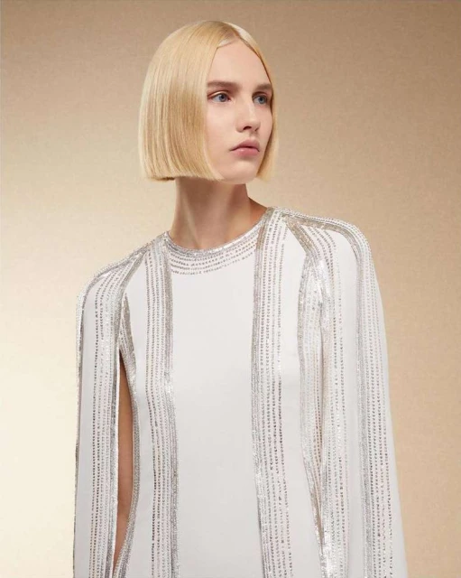 Blonde woman with a bob hairstyle poses in a studio wearing a white dress with silver beaded trim against a beige background.