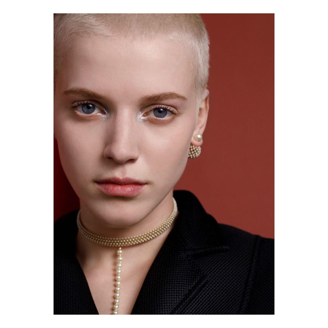Close-up portrait of a young adult female with a buzzcut wearing pearl jewelry and a dark blazer against a terracotta background.