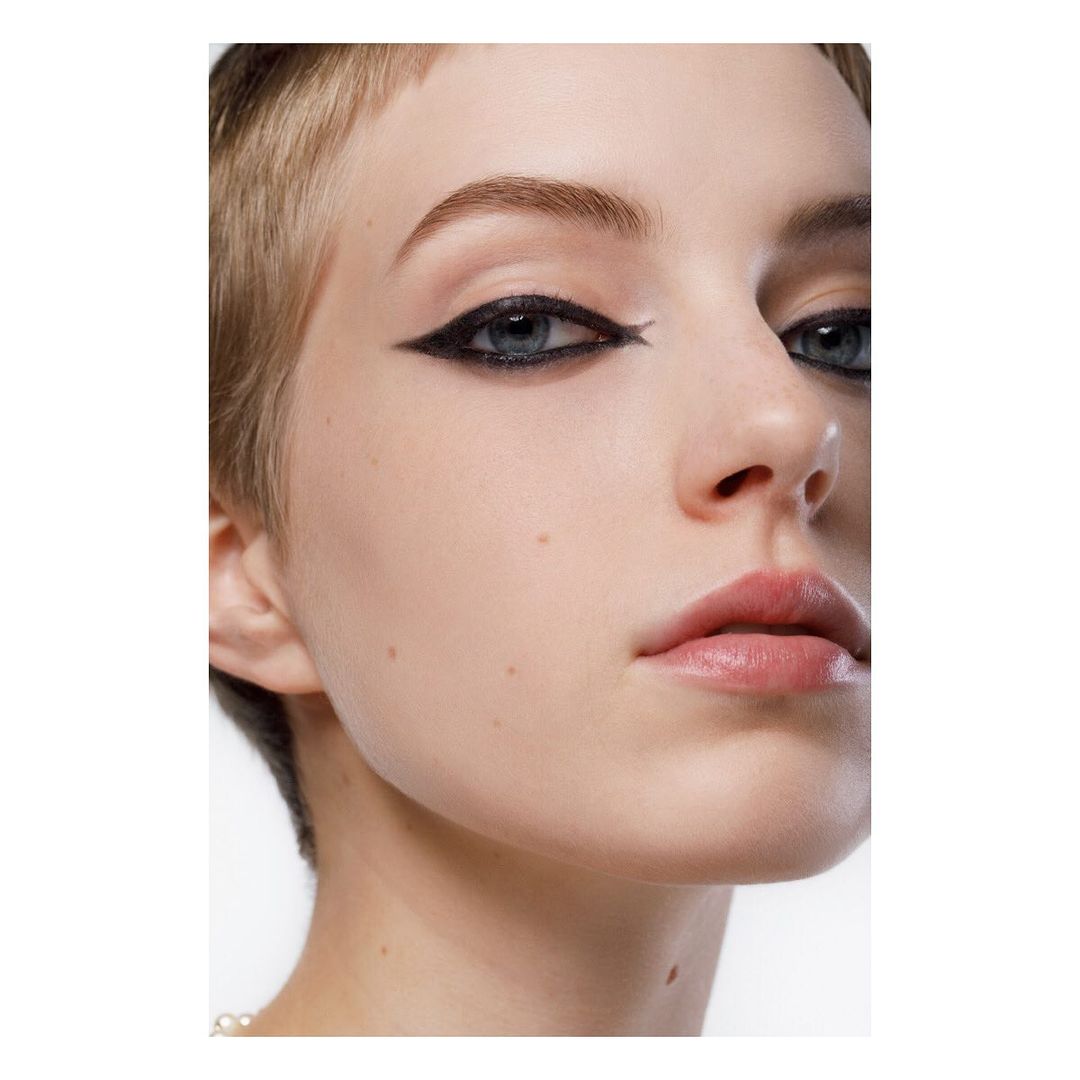 Close-up beauty shot of a fair-skinned woman with a pixie haircut and dramatic winged eyeliner in a studio setting with soft lighting.