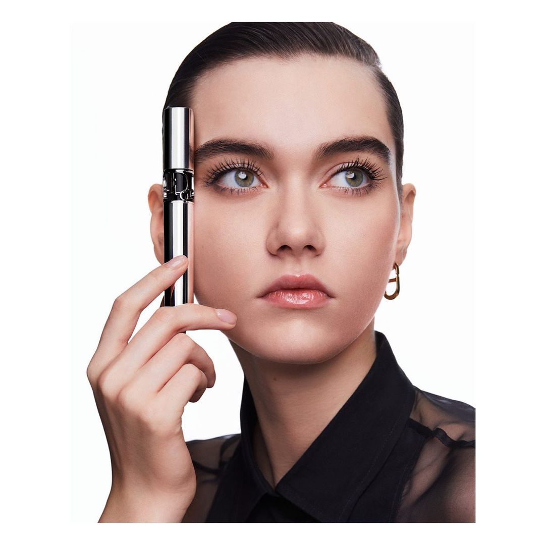 Close-up of a female model holding silver mascara tube. Focus on the model's face, eye makeup, and clean beauty aesthetic.