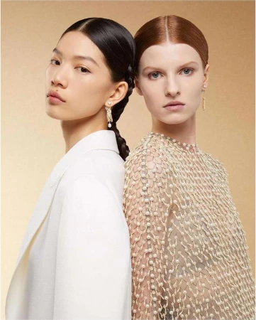 Two female models standing back-to-back against a neutral textured background, wearing white attire and pearl jewelry.