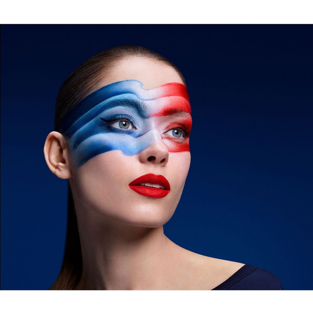 Woman with bold makeup in red, white, and blue, dramatic eyeliner, and red lipstick poses against a dark blue background.