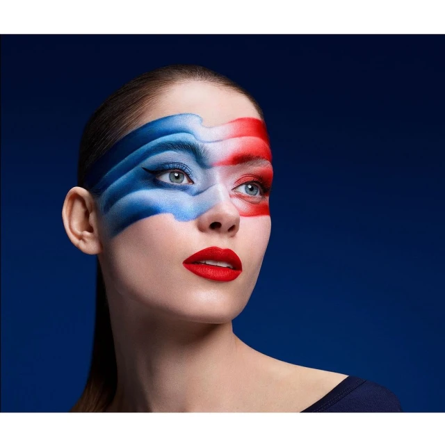 Woman with bold makeup in red, white, and blue, dramatic eyeliner, and red lipstick poses against a dark blue background.