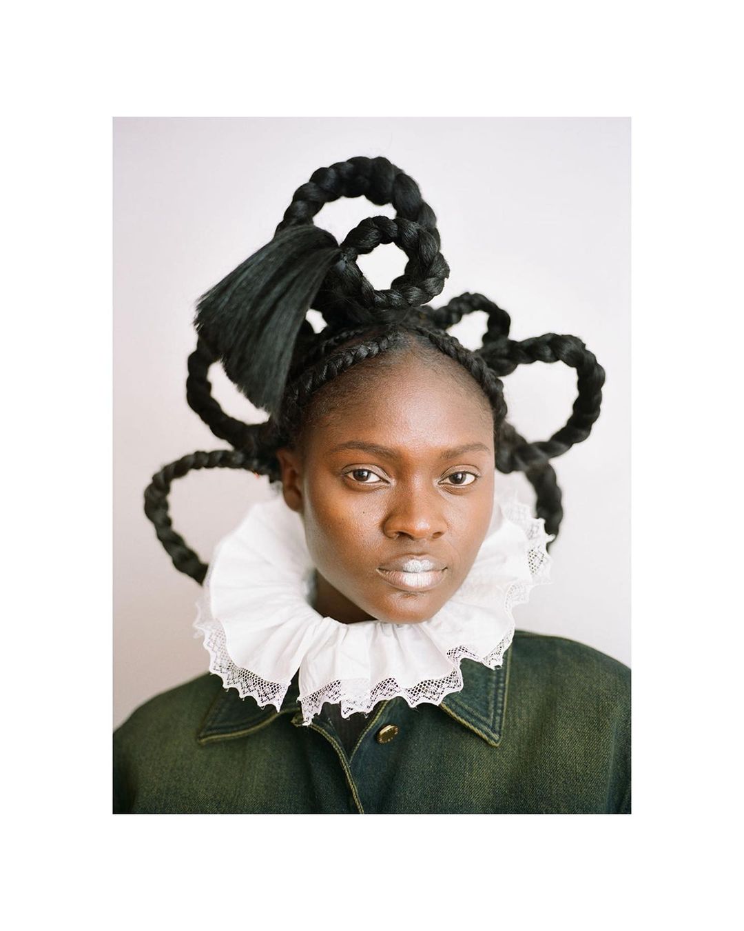 Close-up portrait of an African woman with intricate braided black hairstyle, a white lace ruff collar, and a green denim jacket.