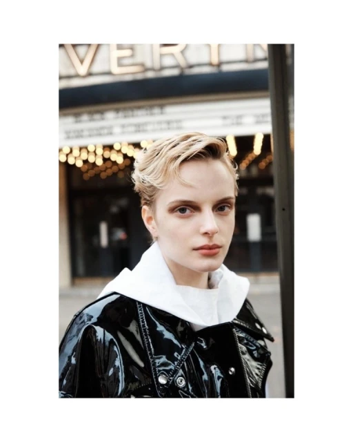 Young woman with a short blonde haircut, wearing a shiny black coat and a white hooded sweatshirt, standing in front of a theater.
