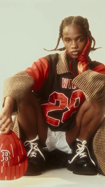 Fashion portrait of a young adult in a baseball jersey, coat, gloves, hat and sneakers, posing in a studio setting with neutral backdrop.