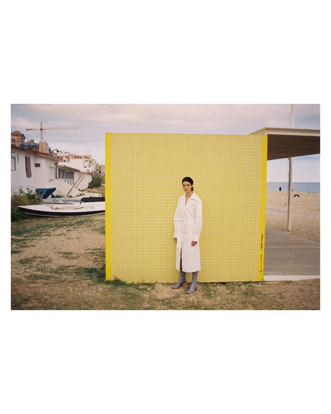 A woman in a trench coat poses near a yellow grid wall by a sandy beach with a coastal town and cloudy sky in the background.