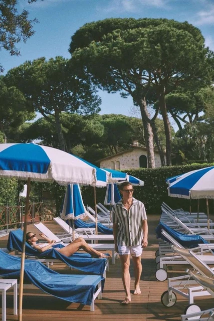 Man walks on a wooden deck near lounge chairs and blue umbrellas on a sunny day, with a woman relaxing, surrounded by lush trees.